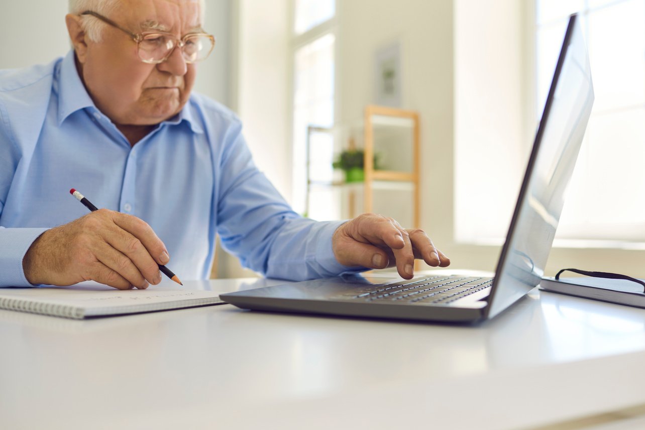 Senior Man in Glasses Using Laptop and Taking Notes in Notebook Sitting at Desk at Home